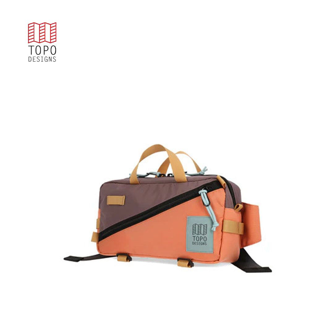 A brown and orange waist pack with a black zipper, branded with 'TOPO DESIGNS', featuring multiple compartments and bottom compression straps.